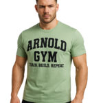 Young man wearing a pistachio green Arnold Gym T-shirt featuring Train. Build. Repeat. slogan, photographed in fitness studio setting.